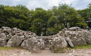 Steingrab aus großen Felsen mit schmalem Eingang und runder Mauer, umgeben von grünen Bäumen unter bewölktem Himmel