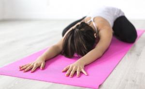 Eine Frau übt auf eine pinken Yogamatte die Stellung des KIndes, mit ausgestreckten Armen und Stirn auf dem Boden.