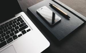 Ein Laptop, ein Stift, ein Smartphone und ein Notizbuch liegen auf einem Schreibtisch.  