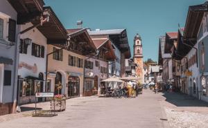 Sonnige Fußgängerzone in Mittenwald mit traditionellen alpenländischen Häusern, Cafés und einer barocken Kirche im Hintergrund.