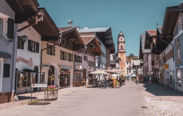 Sonnige Fußgängerzone in Mittenwald mit traditionellen alpenländischen Häusern, Cafés und einer barocken Kirche im Hintergrund.