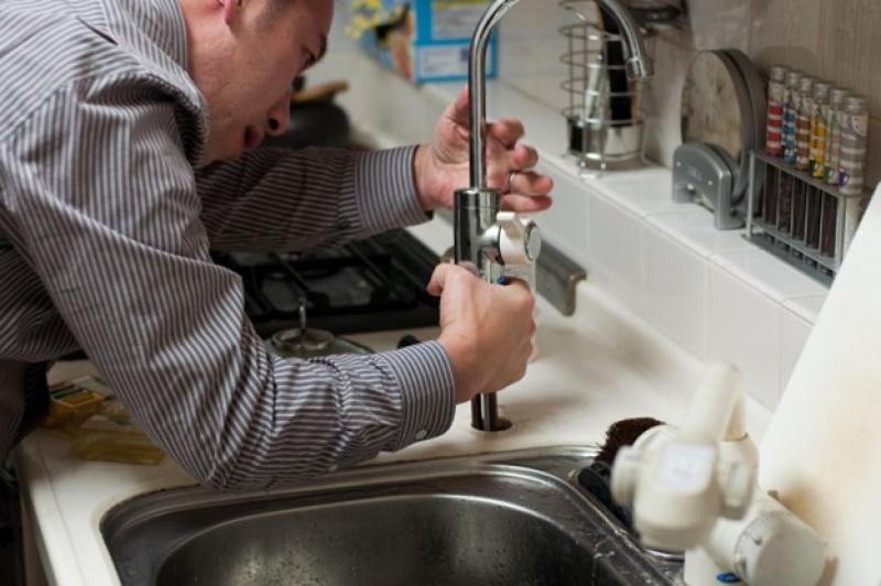 Ein Mann steht in der Küche und repariert den Wasserhahn am Spülbecken.