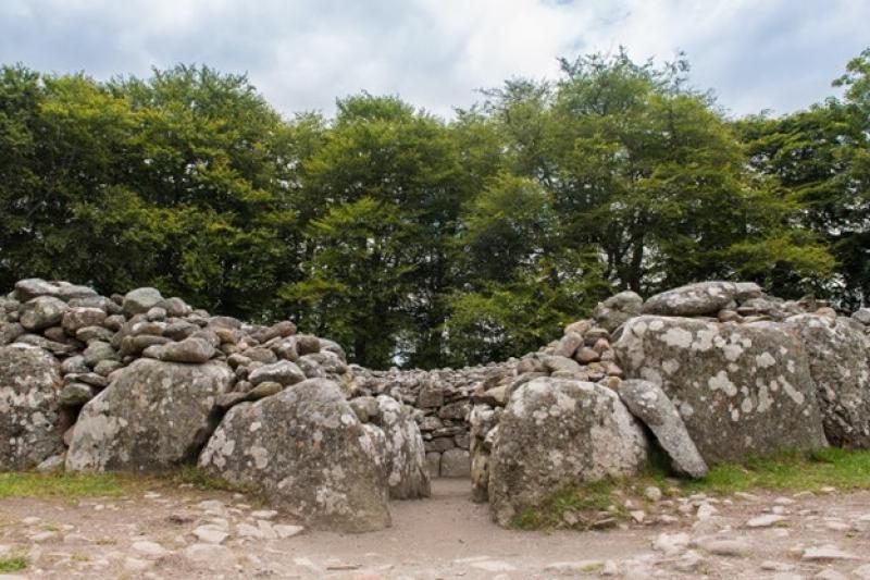 Steingrab aus großen Felsen mit schmalem Eingang und runder Mauer, umgeben von grünen Bäumen unter bewölktem Himmel
