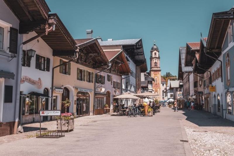 Sonnige Fußgängerzone in Mittenwald mit traditionellen alpenländischen Häusern, Cafés und einer barocken Kirche im Hintergrund.