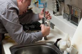Ein Mann steht in der Küche und repariert den Wasserhahn am Spülbecken.