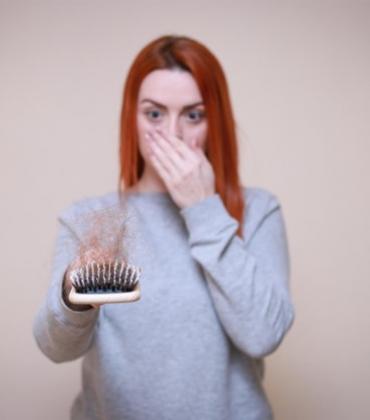 Eine Frau hält eine Bürste mit ausgefallenen Haaren in die Kamera und bedeckt erschrocken ihren Mund.