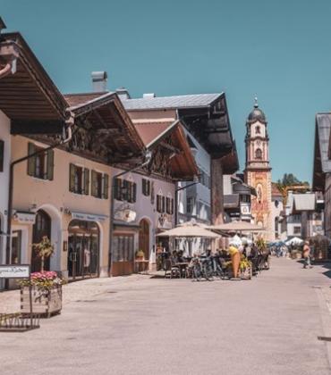 Sonnige Fußgängerzone in Mittenwald mit traditionellen alpenländischen Häusern, Cafés und einer barocken Kirche im Hintergrund.